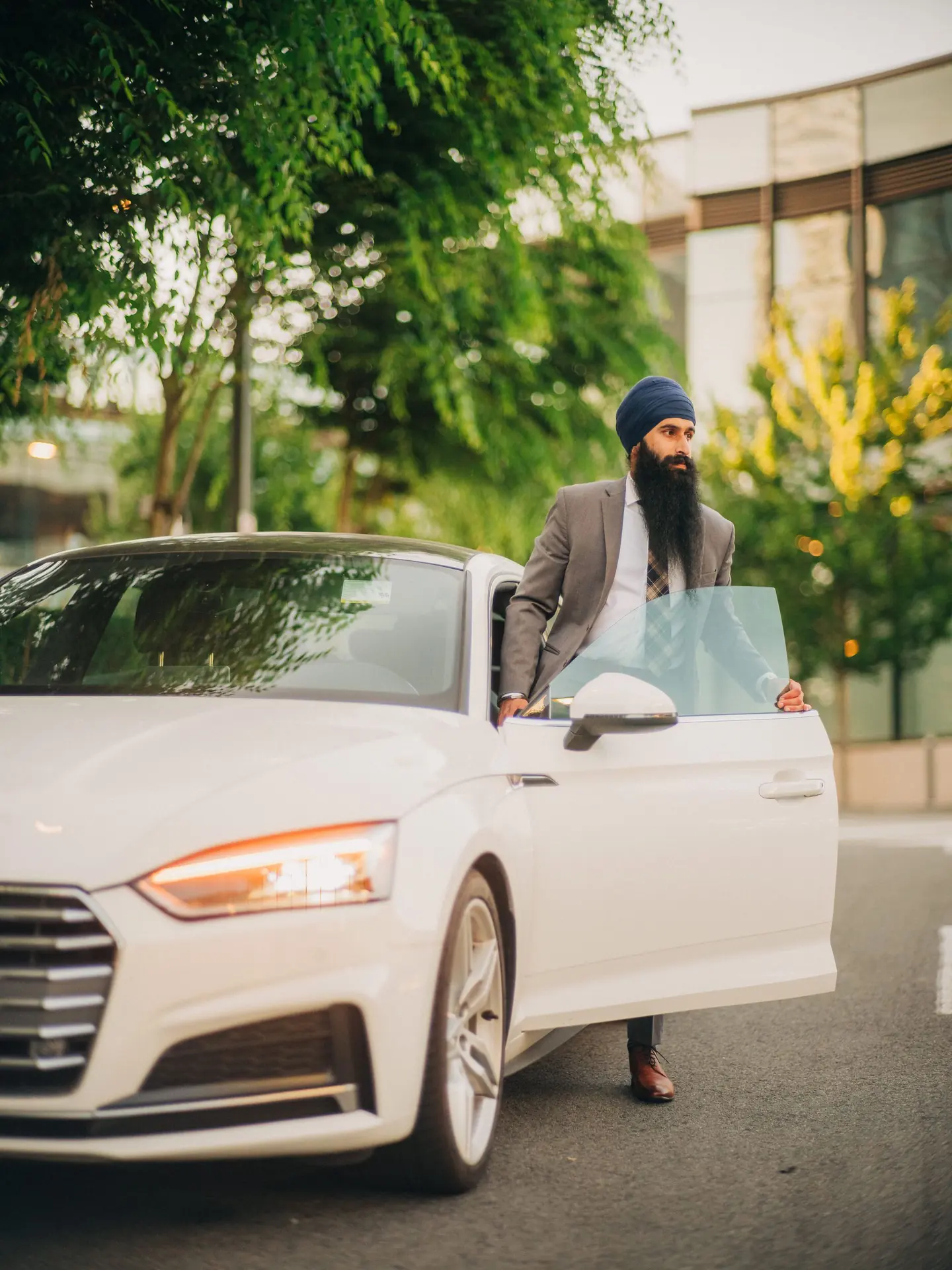 A man in a grey suit and navy turban standing at the open door of a white convertible on a tree lined street.