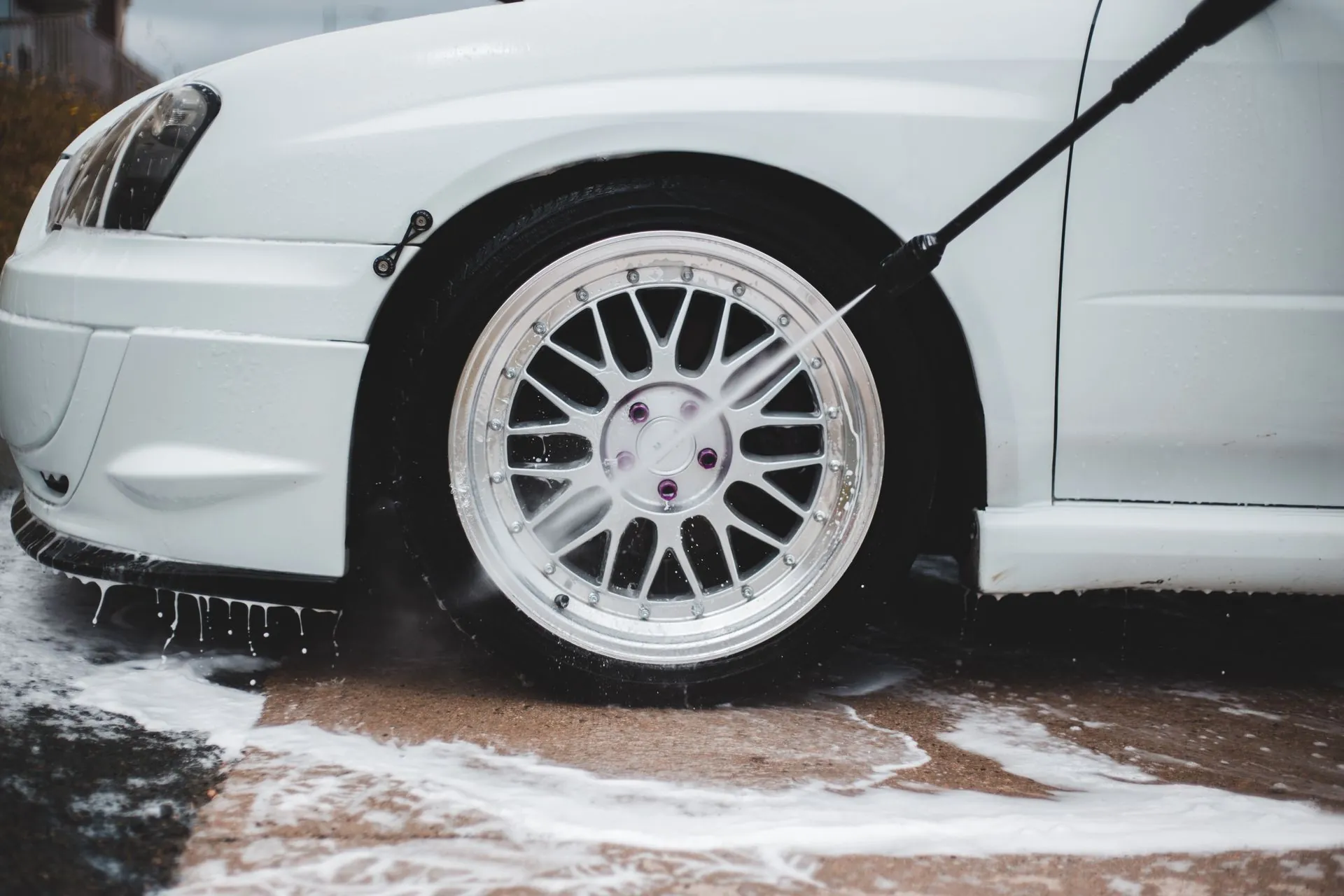 A pressure lance rinsing the front wheel and arch of a white car, foam running off across the ground.